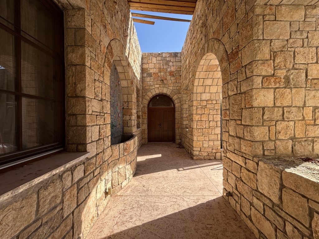 חצר פנימית עם קשתות אבן, דלת עץ מקושתת ומזרקת פסיפס – בית האבן במשמר איילון