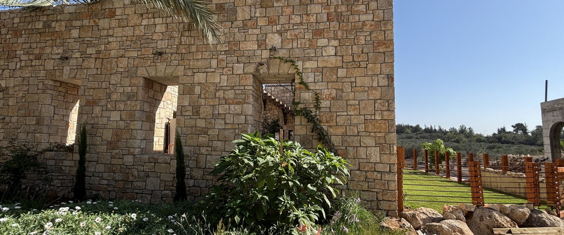 פינת מבנה האבן עם דשא, גדר עץ ונוף פתוח – בית האבן במשמר איילון